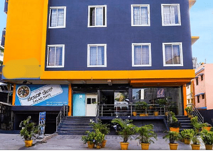 A multi-story building with a blue and yellow facade, visible windows, and potted plants at the entrance | Astra Hotels and Suites Electronic City